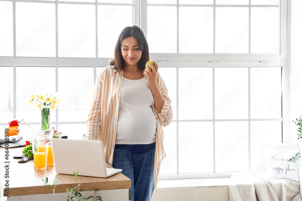 Young pregnant woman with apple using laptop in kitchen