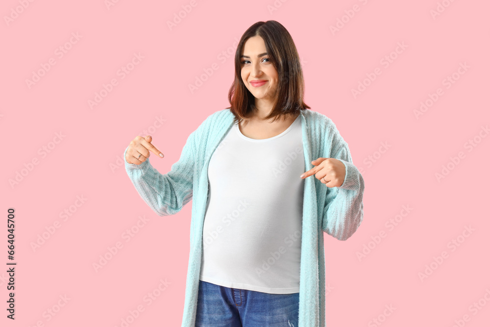 Young pregnant woman pointing at her belly on pink background