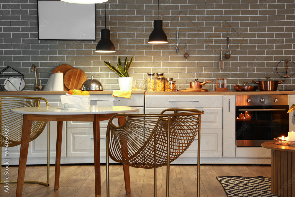 Interior of modern kitchen with glowing lamps, dining table and white counters at evening