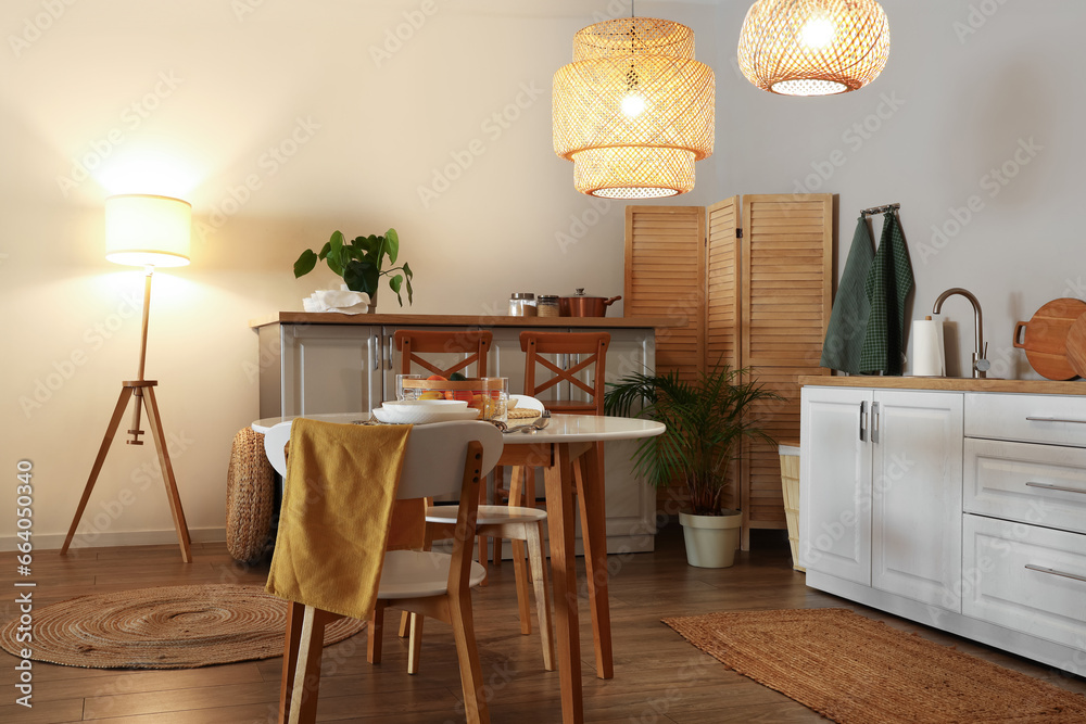 Interior of modern kitchen with glowing lamps, dining table and white counters at evening