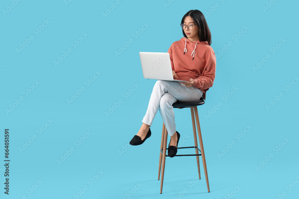 Young female Asian programmer with laptop sitting on chair against blue background