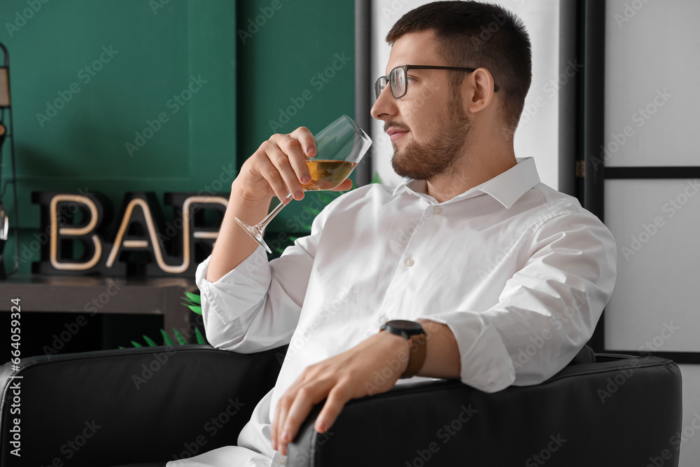 Young sommelier with glass of wine sitting on black armchair in living room