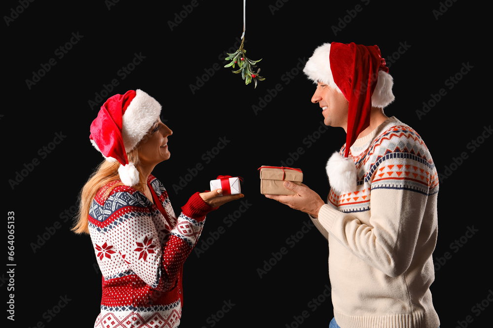 Mature couple with Christmas gifts kissing under mistletoe branch on dark background