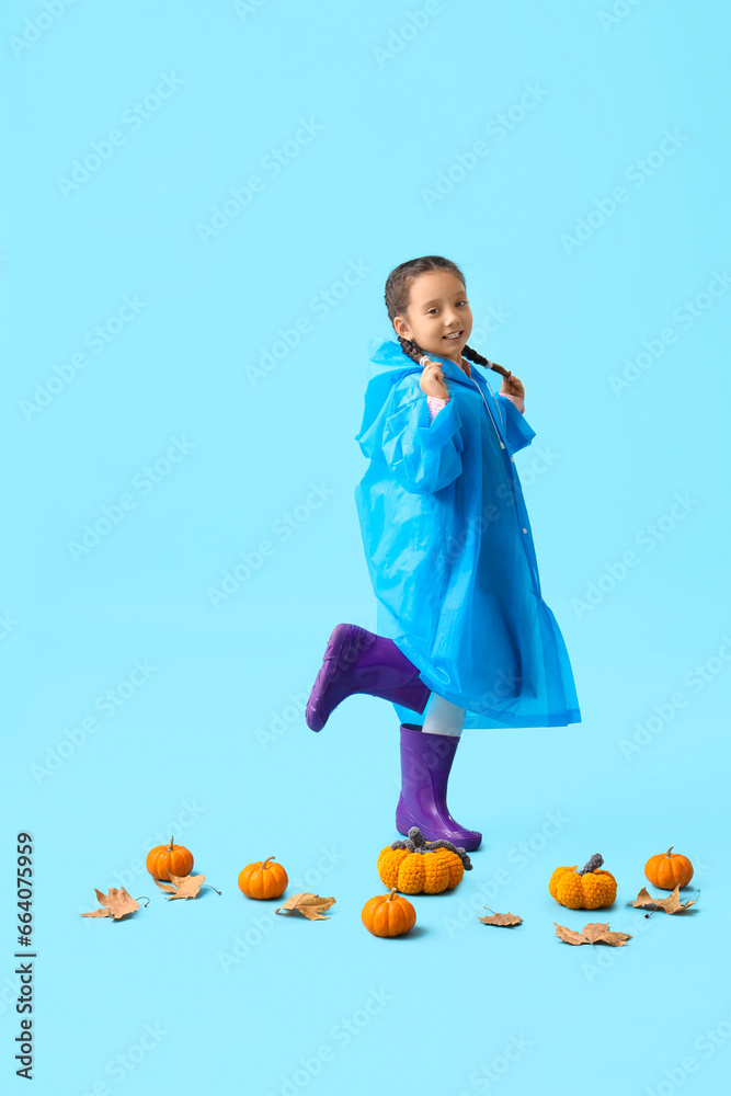 Little Asian girl in rubber boots with pumpkins on blue background