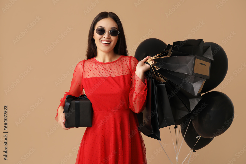 Beautiful young Asian woman with shopping bags, gift box and balloons on brown background. Black Friday sale