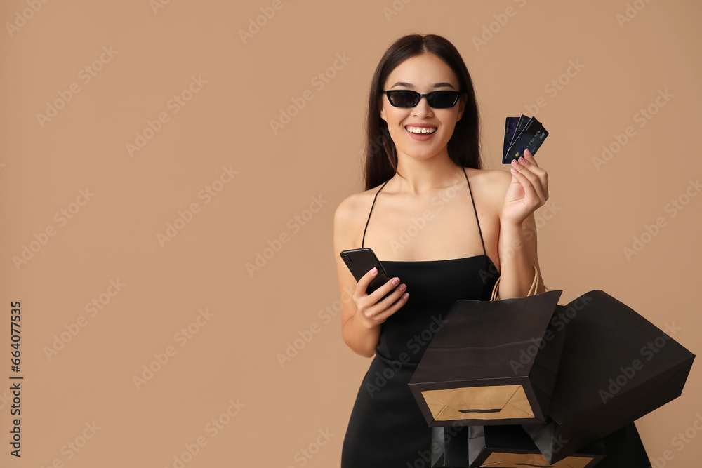 Beautiful young Asian woman with shopping bags, mobile phone and credit cards on brown background. Black Friday sale