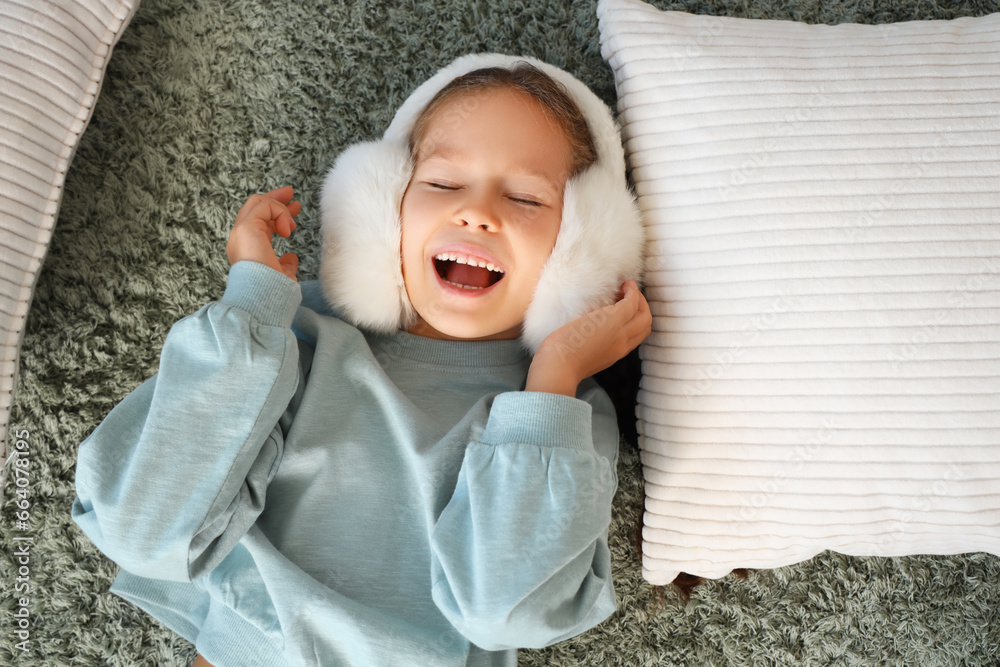 Cute little girl in warm earmuffs lying on carpet at home, top view