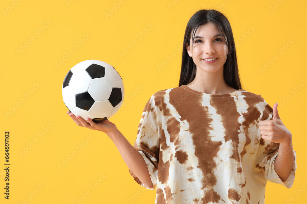 Young woman with soccer ball showing thumb-up on yellow background