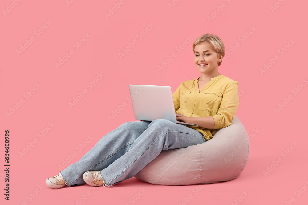 Happy young female programmer with laptop sitting on beanbag chair against pink background