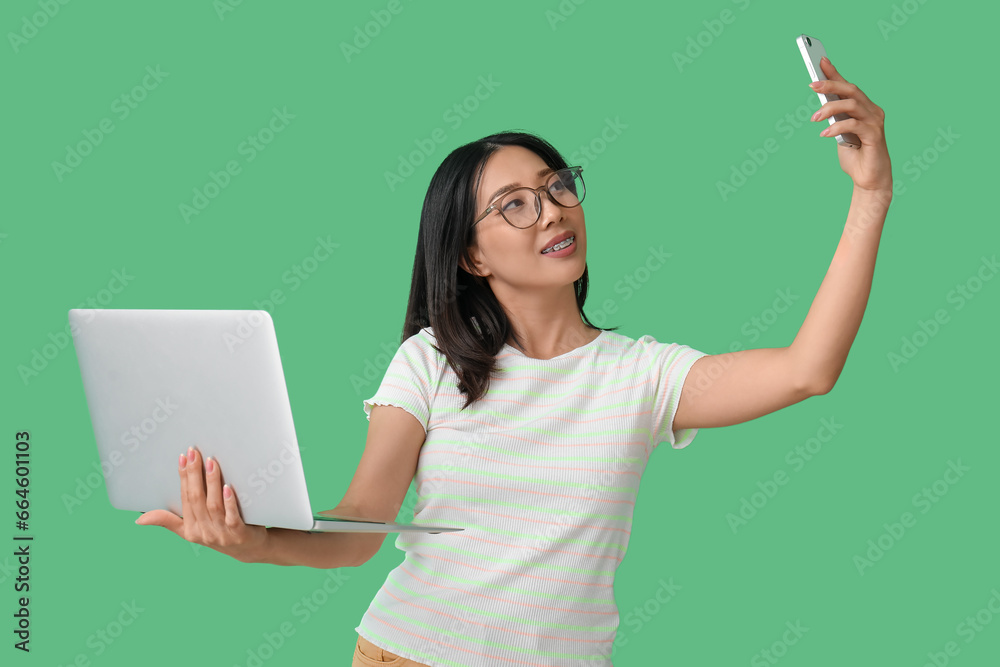 Happy young female Asian programmer in eyeglasses with laptop taking selfie on green background