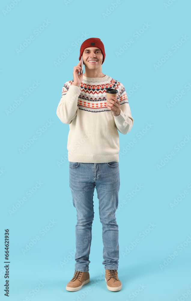 Young man in winter clothes with cup of coffee talking by mobile phone on blue background