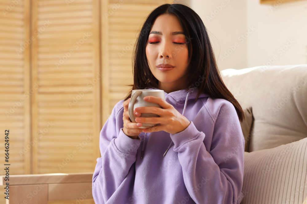 Young Asian woman in hoodie with cup of coffee at home