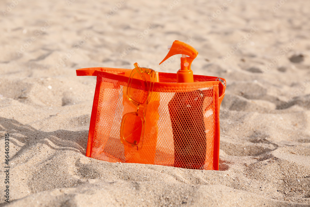 Cosmetic bag with bottles of sunscreen cream and sunglasses on sandy background