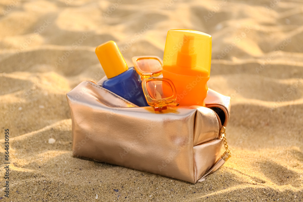 Cosmetic bag with bottles of sunscreen cream and sunglasses on sandy background