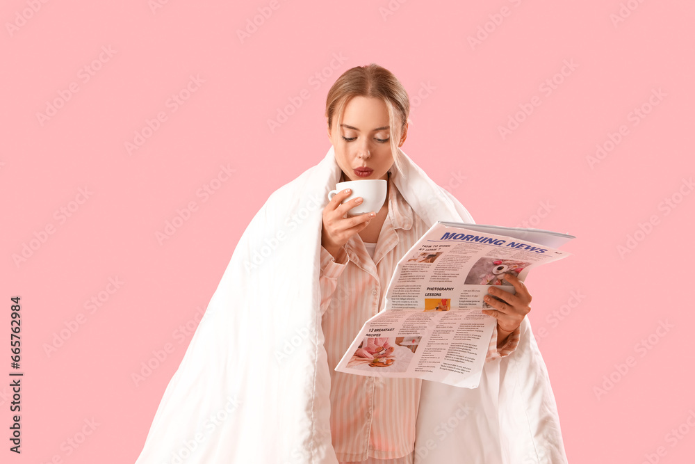 Young woman in pajamas with cup of coffee reading newspaper on pink background