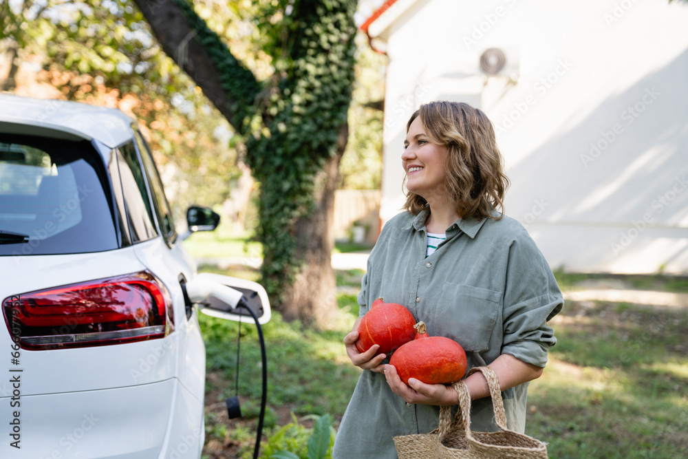 Woman with shopping bag and pumpkins next to a charging electric car in the yard of a country house