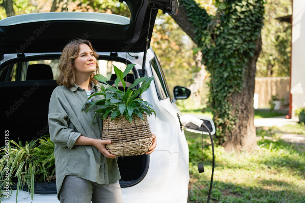 Woman with plant in pot next to a charging electric car in the yard of a country house