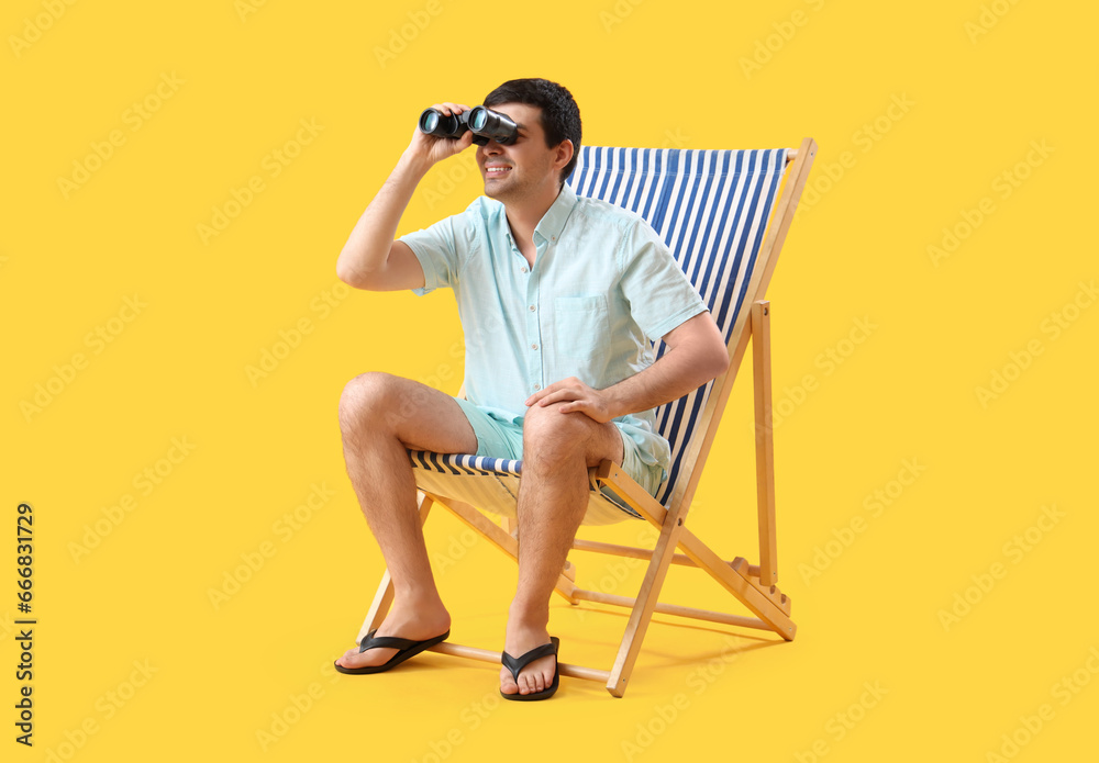 Young man with binoculars sitting in deck chair on yellow background