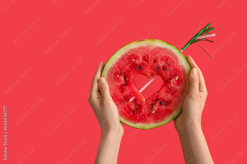 Female hands with half of ripe watermelon, straw and mini umbrella on red background