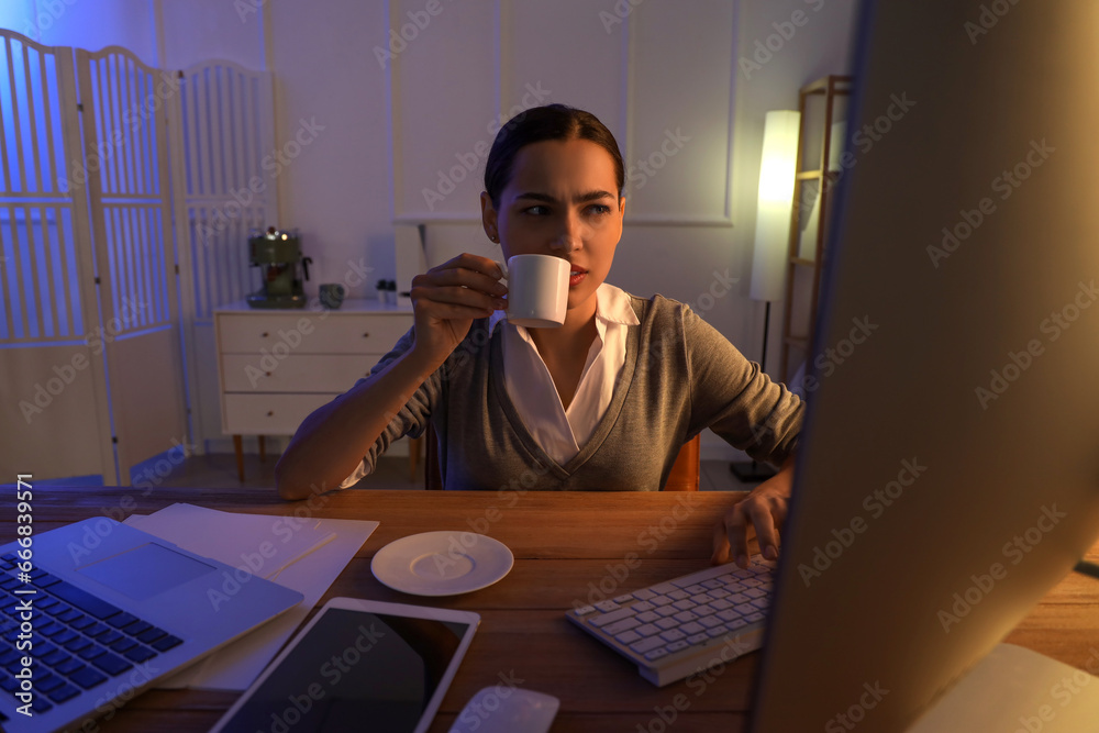 Tired young female programmer with cup of coffee working in office at night