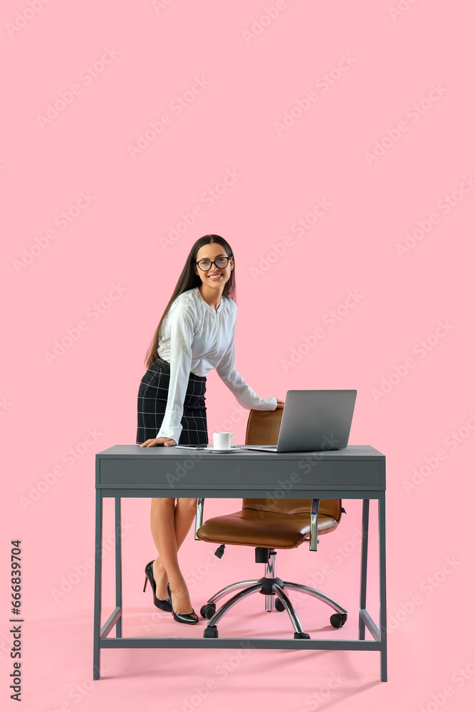 Pretty young female programmer working at table with laptop on pink background