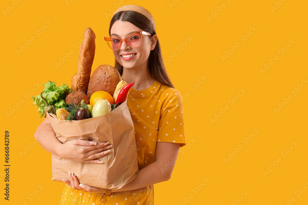 Young woman holding paper bag with fresh vegetables on yellow background