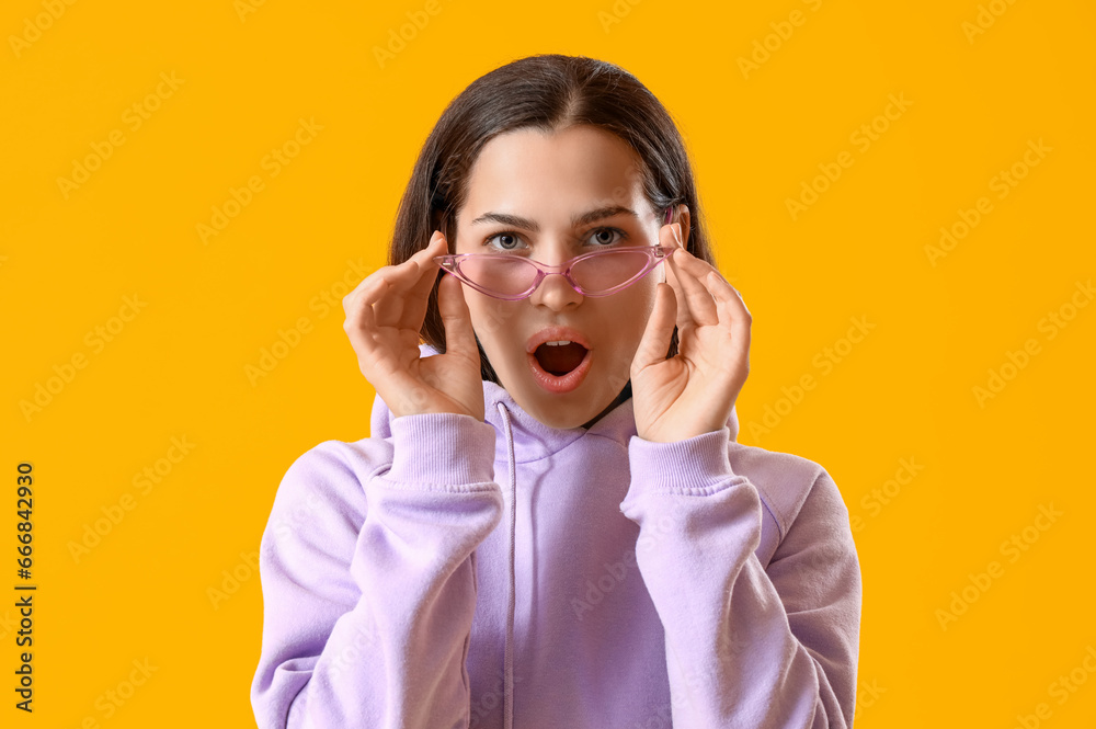 Surprised young woman in lilac hoodie and sunglasses on yellow background, closeup