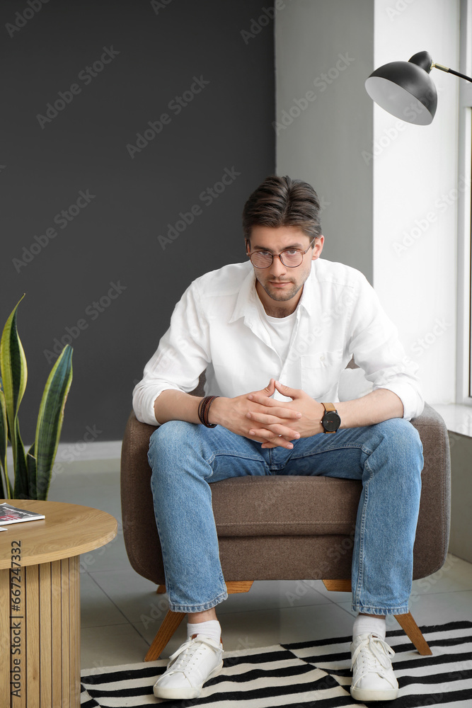 Handsome young man with wristwatch and eyeglasses sitting on armchair in room