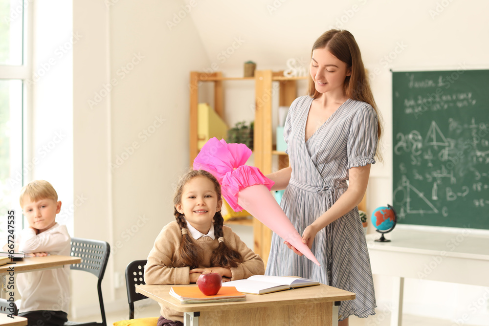 Teacher greeting her happy pupil with pink school cone in classroom