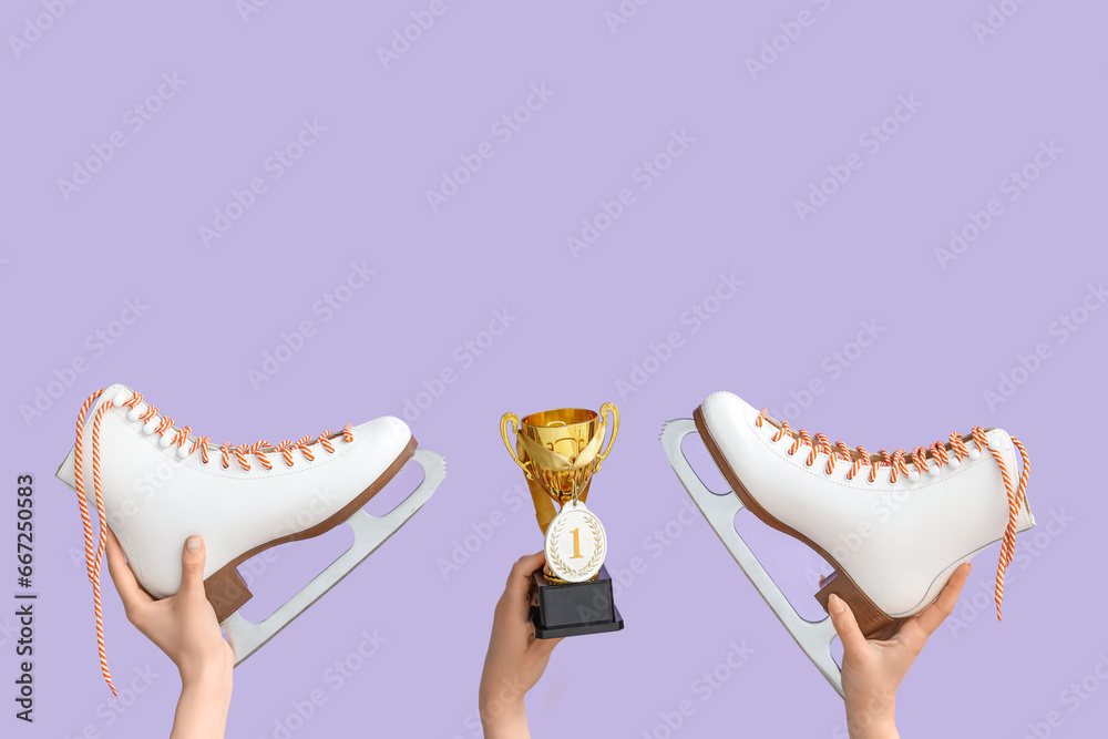 Female hands with gold cup, medal and ice skates on lilac background