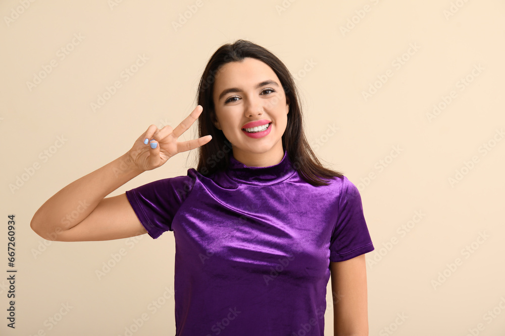 Beautiful happy young woman showing victory gesture on beige background