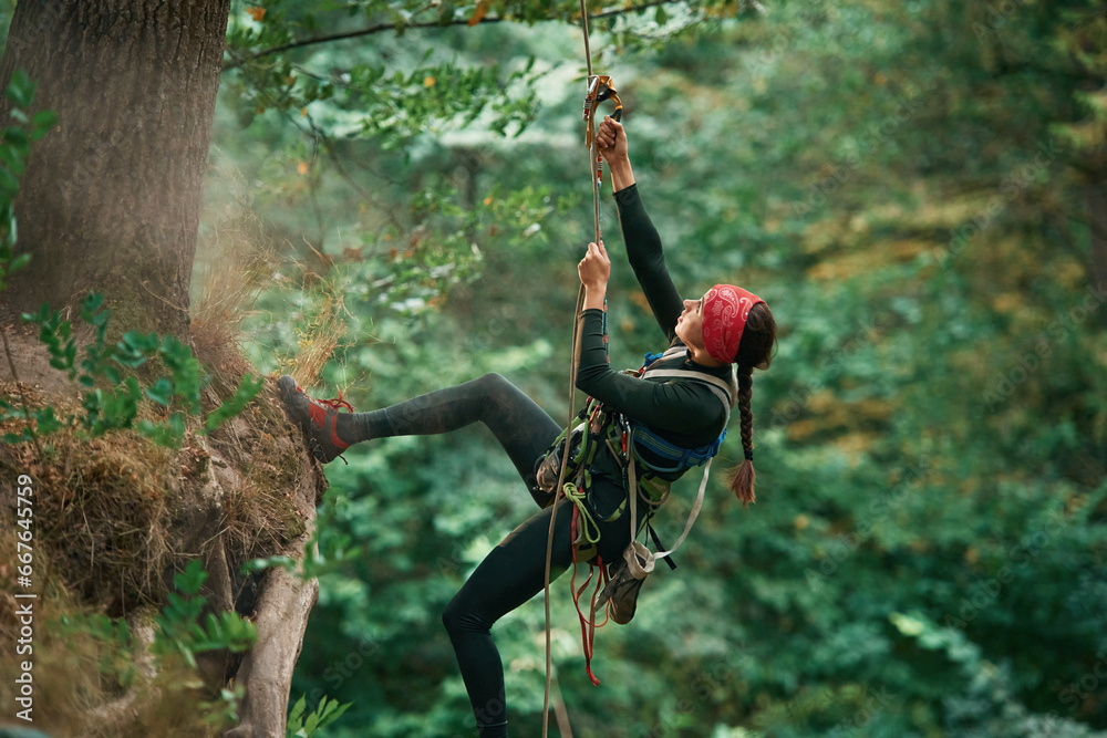 View from the side, hanging on the rope. Woman is doing climbing in the forest by the use of safety equipment