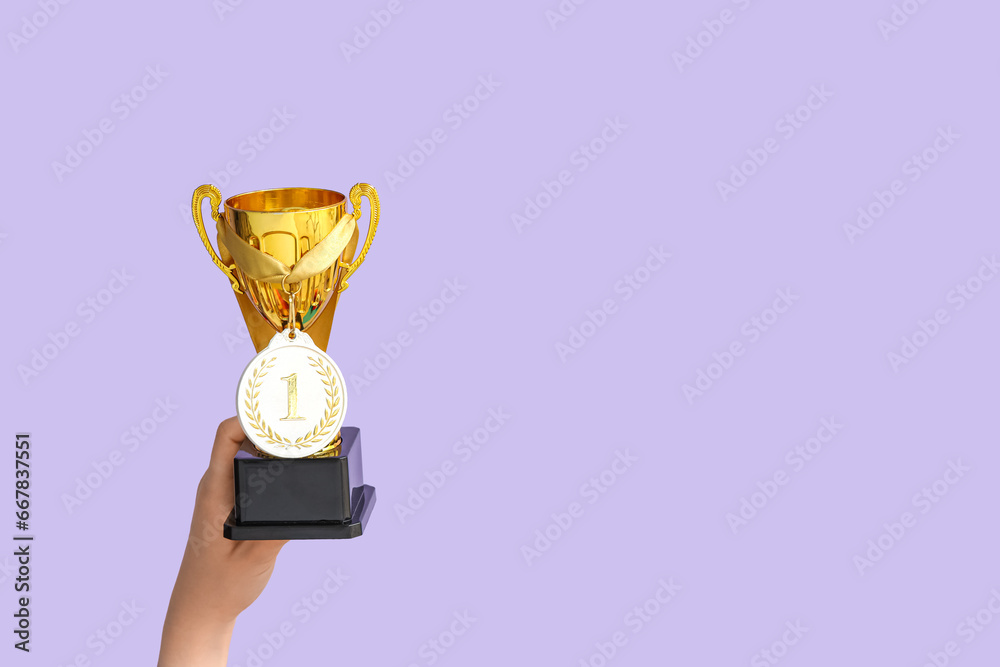 Female hands with gold cup and medal on lilac background