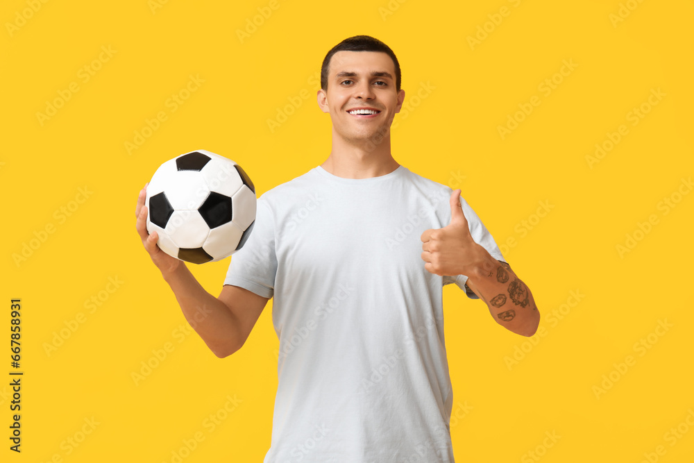 Handsome young man with soccer ball showing thumb-up gesture on yellow background