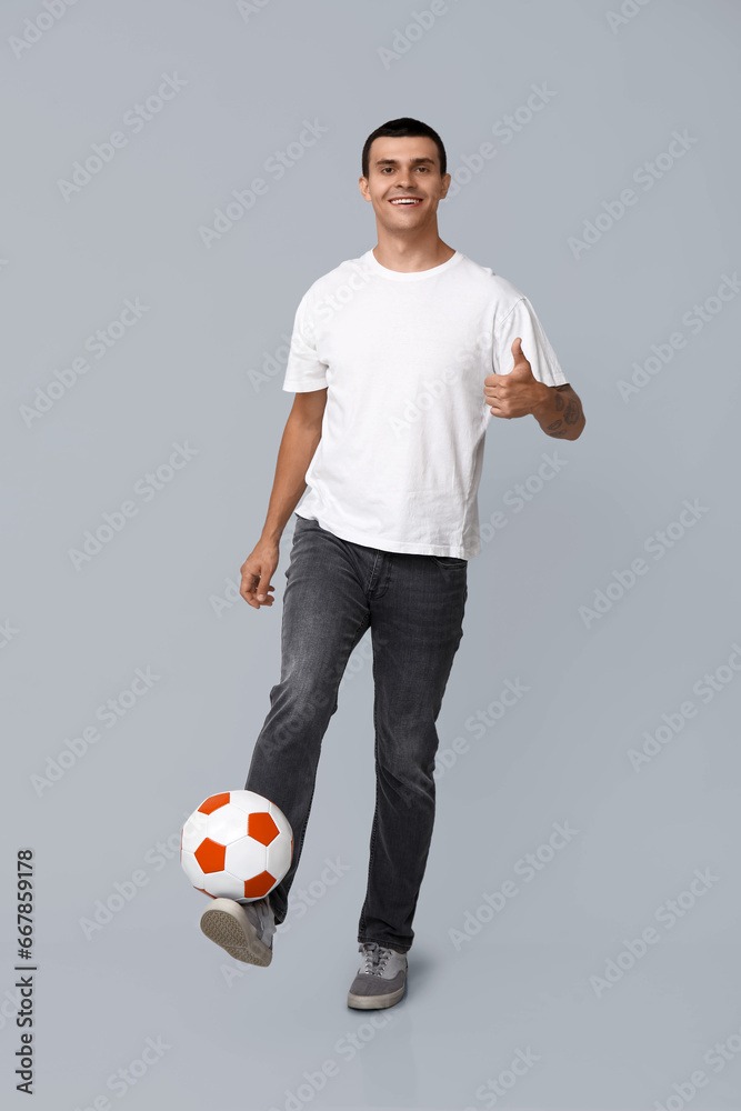 Handsome young man with soccer ball showing thumb-up gesture on grey background