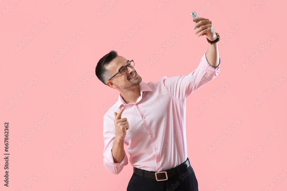 Happy young businessman with mobile phone taking selfie on pink background