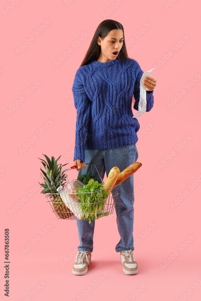 Shocked young woman with shopping basket and receipt from store on pink background