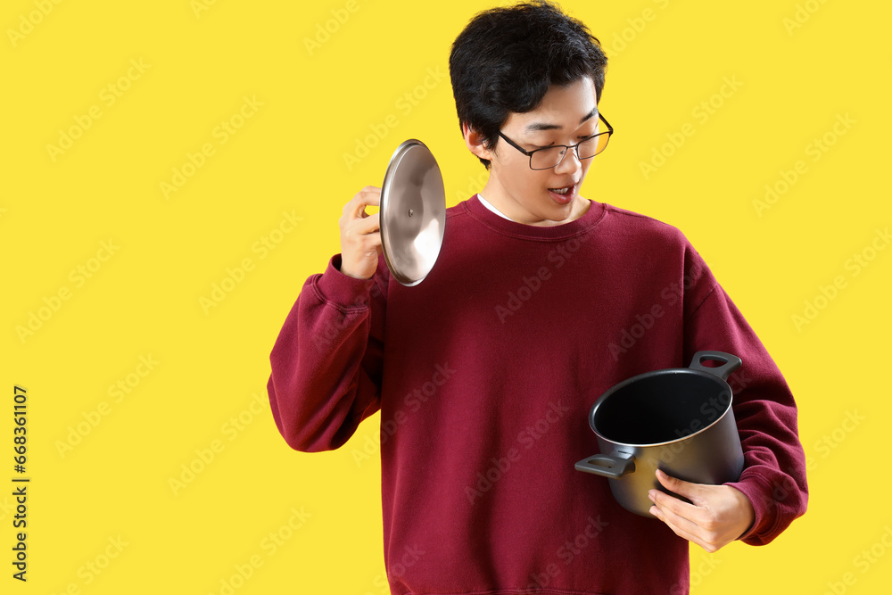 Young Asian man with cooking pot on yellow background