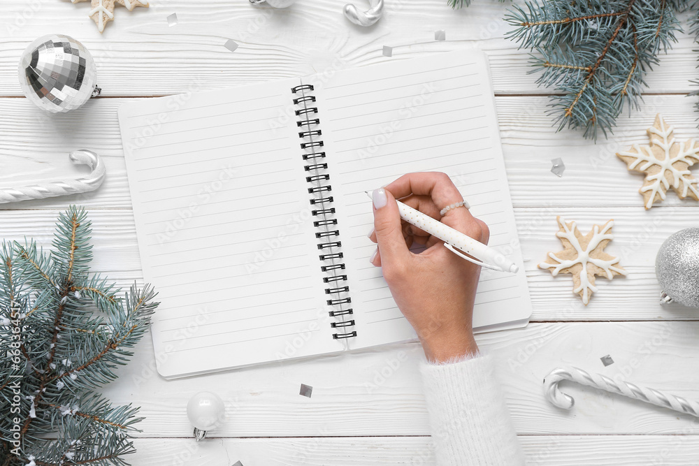 Woman writing in notebook with Christmas decor and cookies on white wooden background