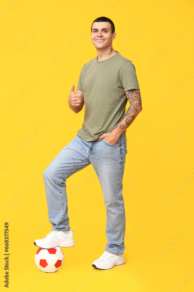 Handsome young man with soccer ball showing thumb-up gesture on yellow background