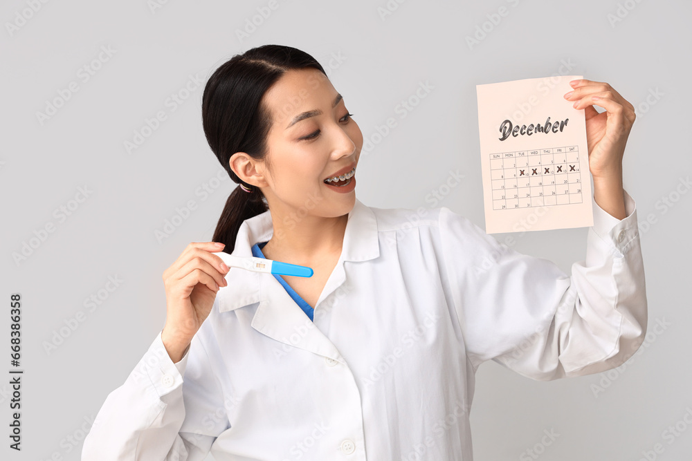 Female Asian doctor with pregnancy test and calendar on light background