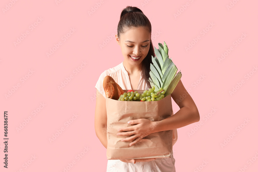 Young Asian woman with shopping bag full of fresh food on pink background