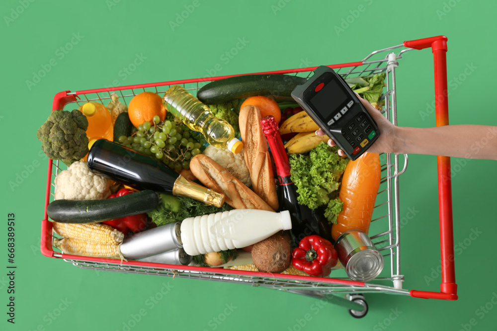 Shopping cart full of food and female hand with payment terminal on green background