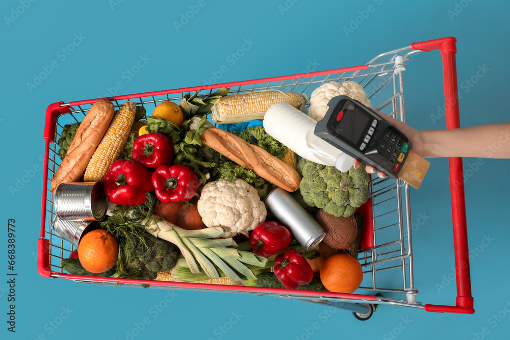 Shopping cart full of food and female hand with payment terminal on blue background