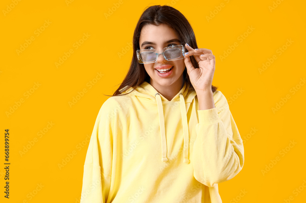 Beautiful young woman in stylish hoodie and sunglasses on yellow background