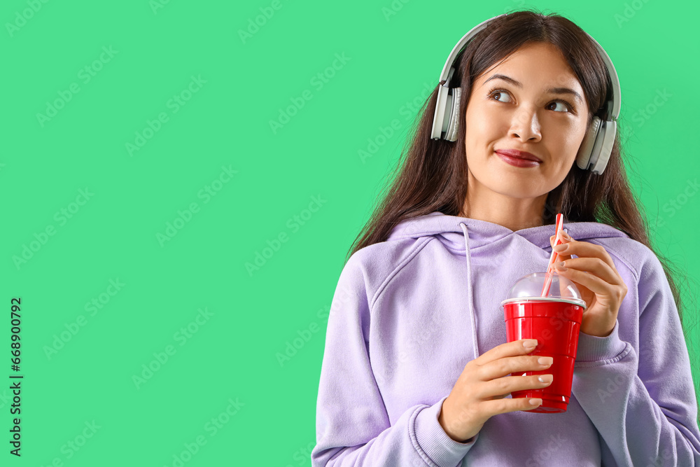 Beautiful young Asian woman in stylish hoodie and headphones holding cup of drink on green background