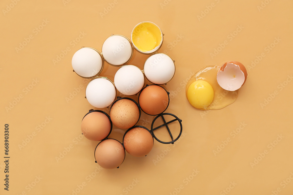 Holders with cracked and whole chicken eggs on beige background