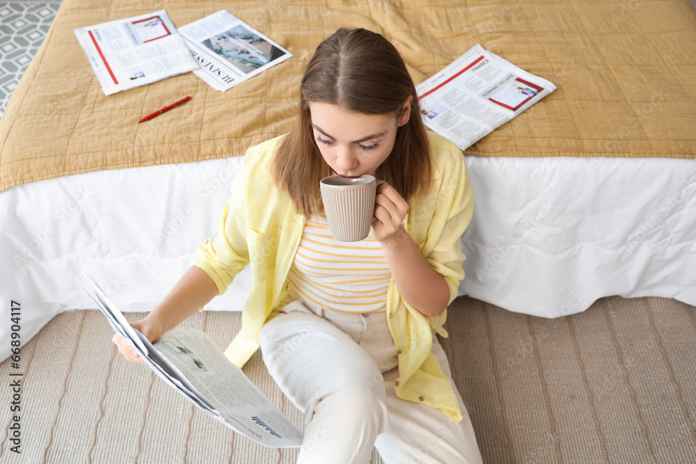 Beautiful young woman with cup of tea reading newspaper in bedroom at home