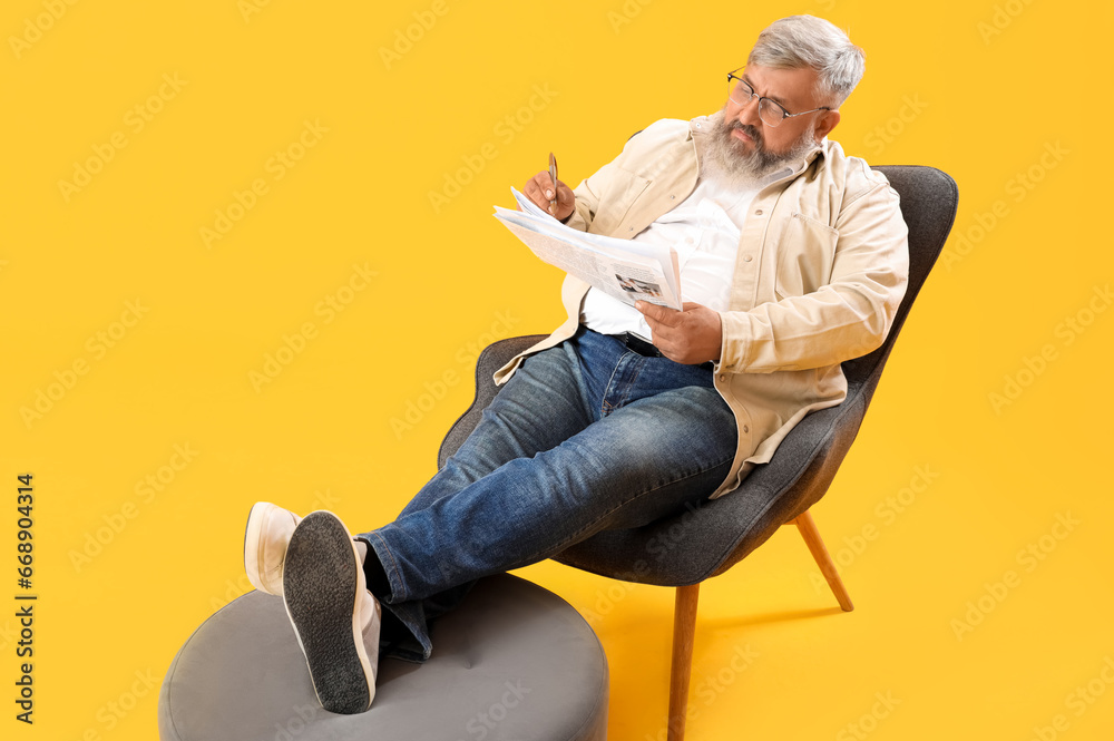 Mature man with pen and newspaper sitting on armchair against yellow background