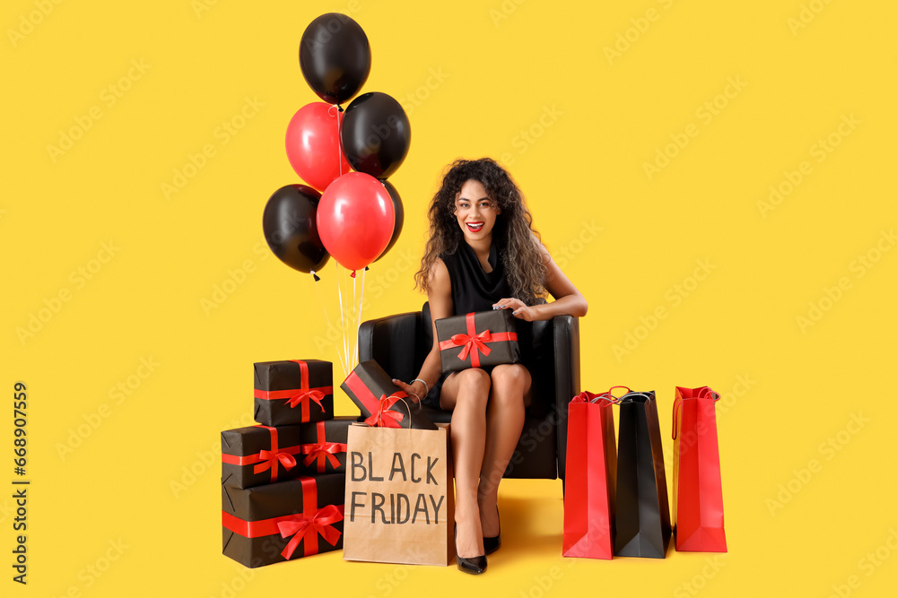 Beautiful African-American woman with shopping bags, gift boxes and balloons sitting on chair against yellow background. Black Friday sale